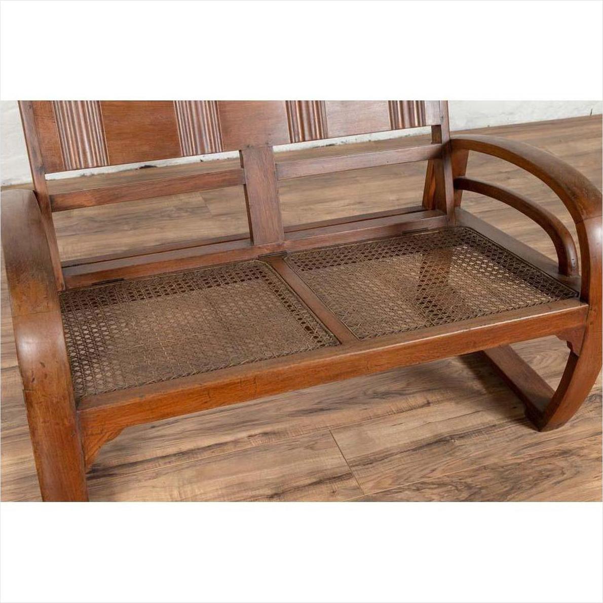 Brown teak wood settee with folding back, looping arms, and cane seat on wooden floor indoors.