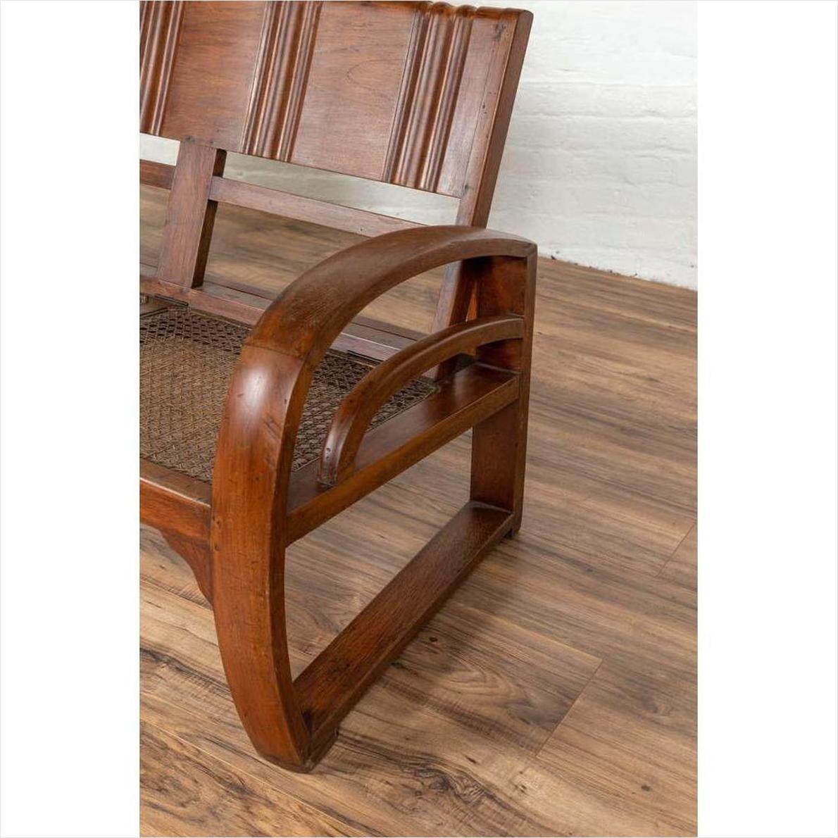 Close-up of brown teak wood settee with cane seat, folding back, and looping arms on a wooden floor.