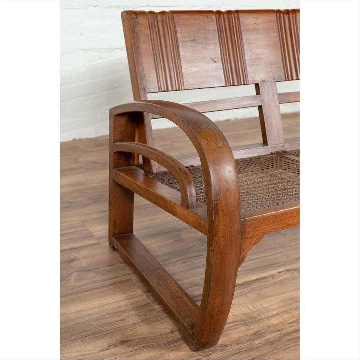 Brown teak wood settee with looping arms, folding back, and cane seat on a wooden floor indoors.