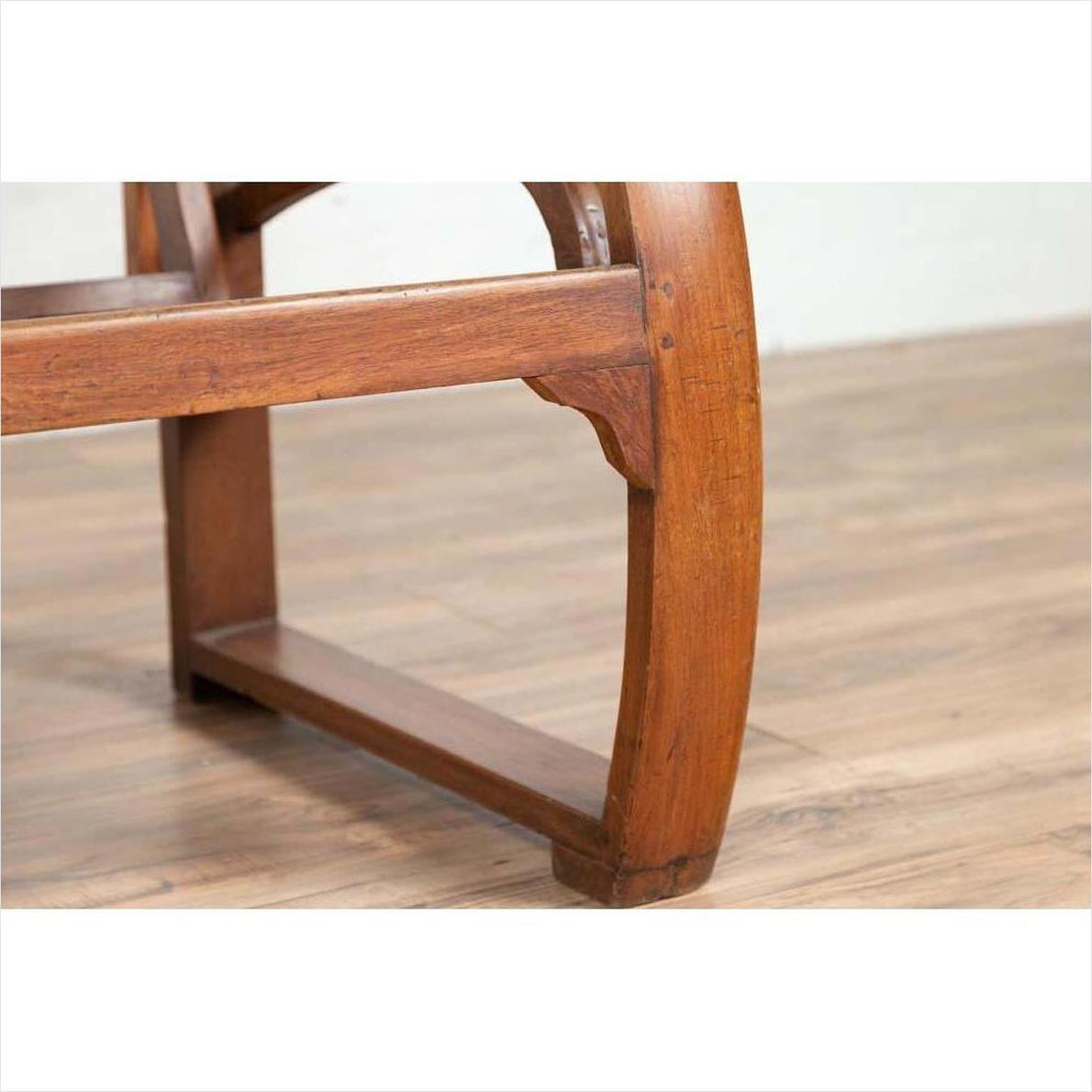 Close-up of brown teak wood settee with looping arms, folding back, and cane seat on a light wood floor indoors.