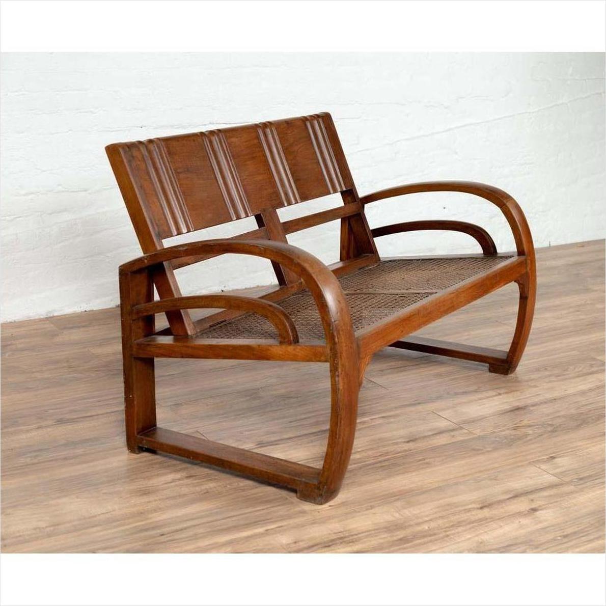 Brown teak wood settee with folding back, looping arms, and cane seat against a white indoor wall.