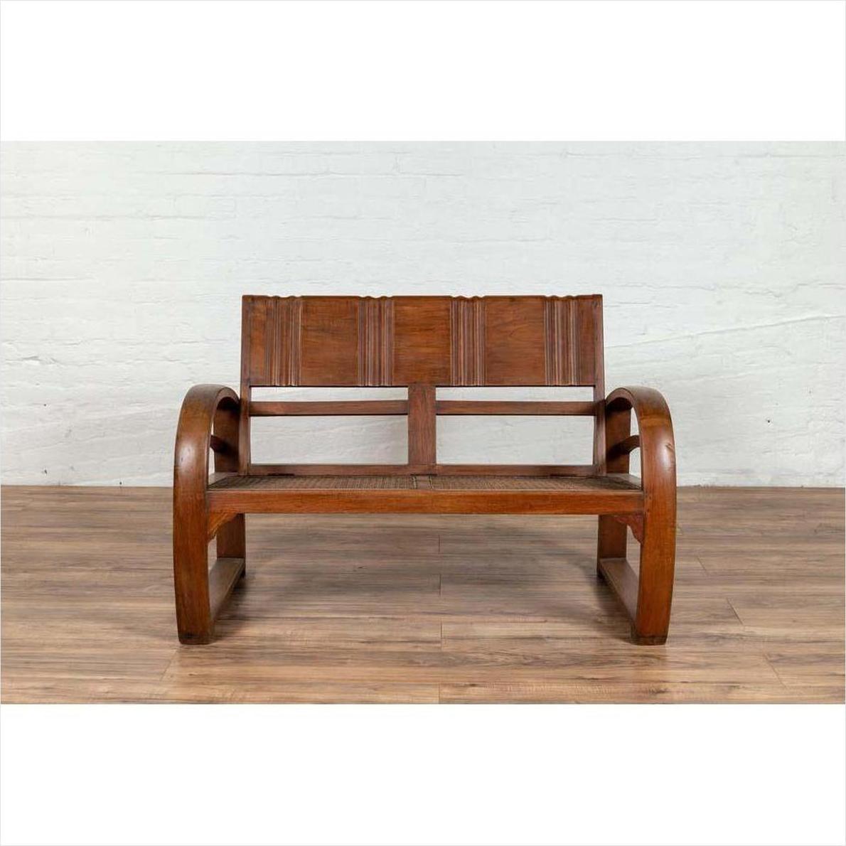 Brown teak wood settee with looping arms, folding back, and cane seat on a wooden floor indoors.