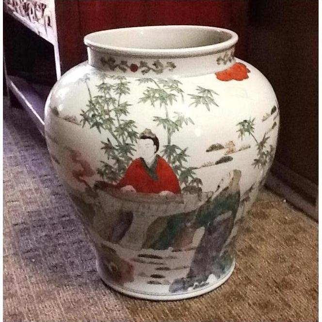 White porcelain urn with painted figures, bamboo, and outdoor scene on a carpeted floor near wooden furniture.