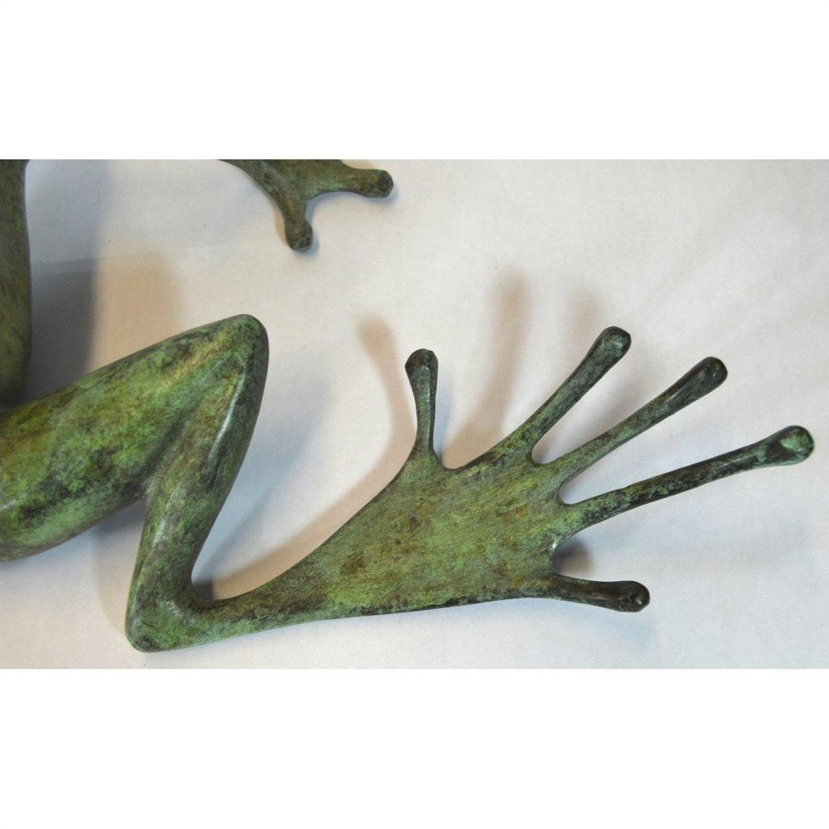 Close-up of greenish bronze frog sculpture with bent leg and webbed foot, light background.