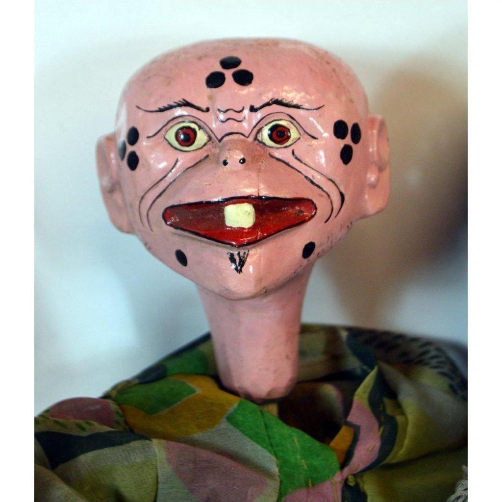 Close-up of vintage Balinese puppet with painted pink face, wide eyes, black spots, big tooth, colorful fabric clothing.