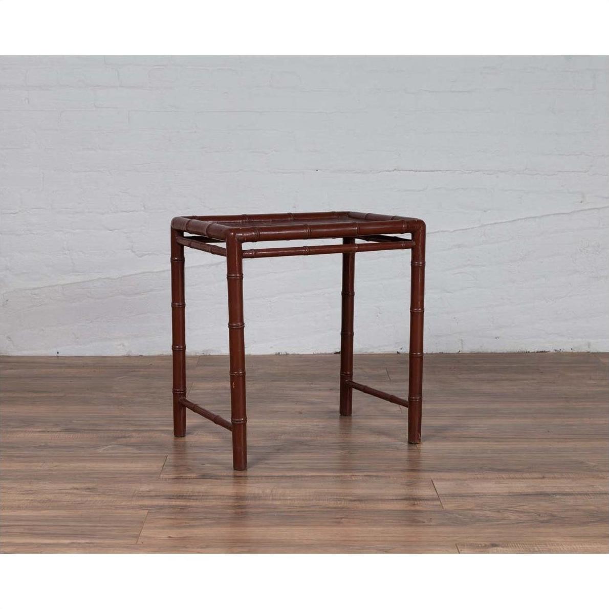 Brown bamboo side table with recessed top, vintage 1950s style, on wooden floor by white brick wall indoors.