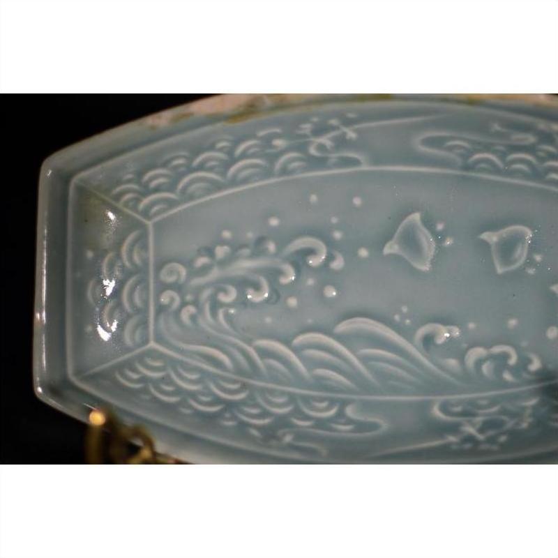 Light blue celadon ceramic dish with embossed wave and fish design, top view against a dark background.