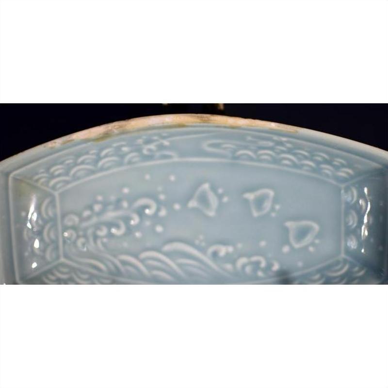 Close-up of light blue ceramic celadon dish with raised wave and fish patterns on the edge.