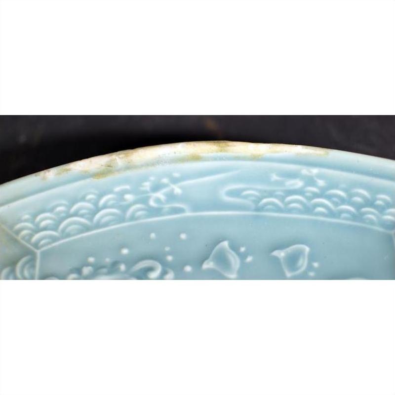 Close-up of a green celadon ceramic dish with embossed patterns and a chipped, weathered rim.