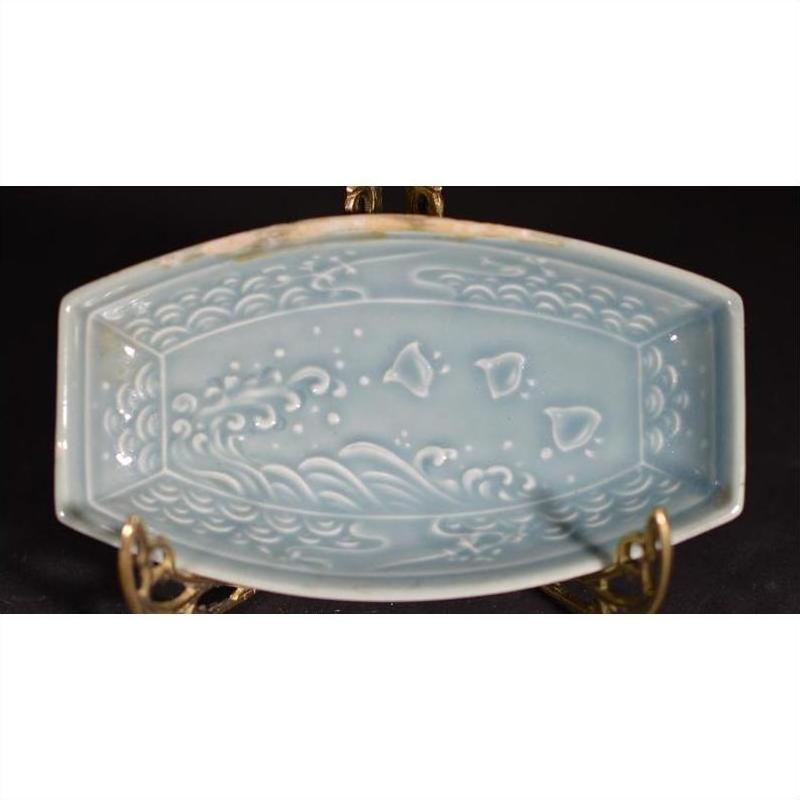 Green celadon ceramic dish with wave and fish patterns on a brass stand against a dark background, close-up.