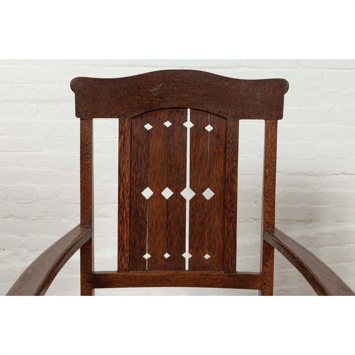 Close-up of brown wood Dutch Colonial armchair with pierced slats and bonnet rail, indoors by white brick wall.
