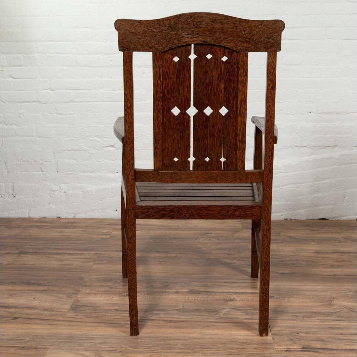 Brown wooden Dutch Colonial armchair with pierced slats and bonnet rail on wooden floor by white brick wall indoors.