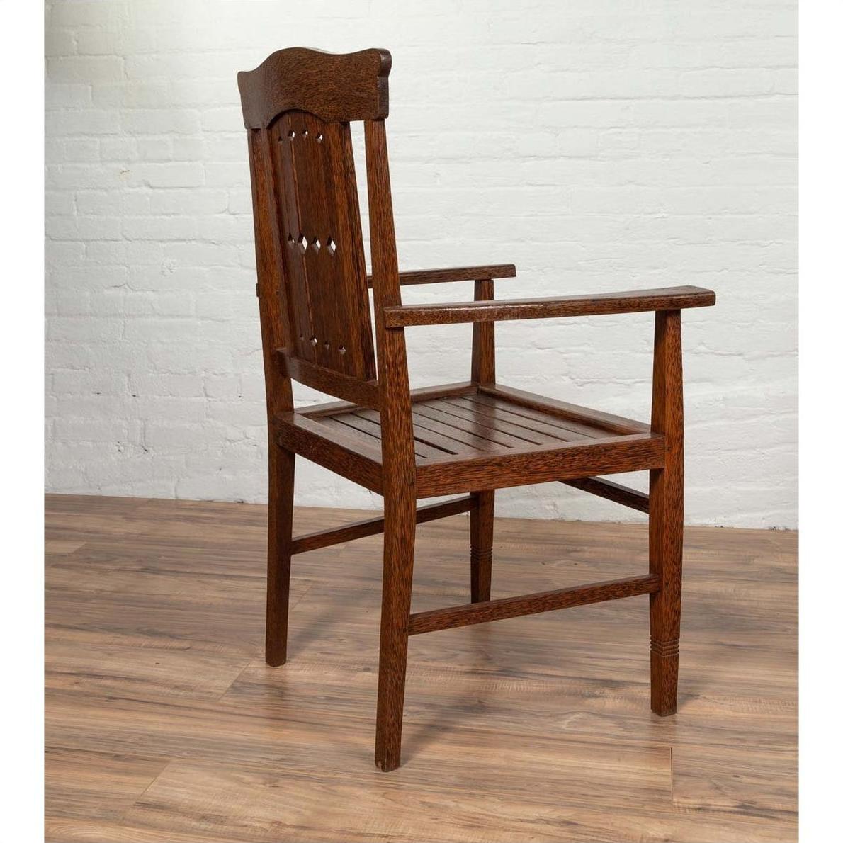 Brown wood Dutch Colonial armchair with pierced slats and bonnet rail on wooden floor near white brick wall indoors.