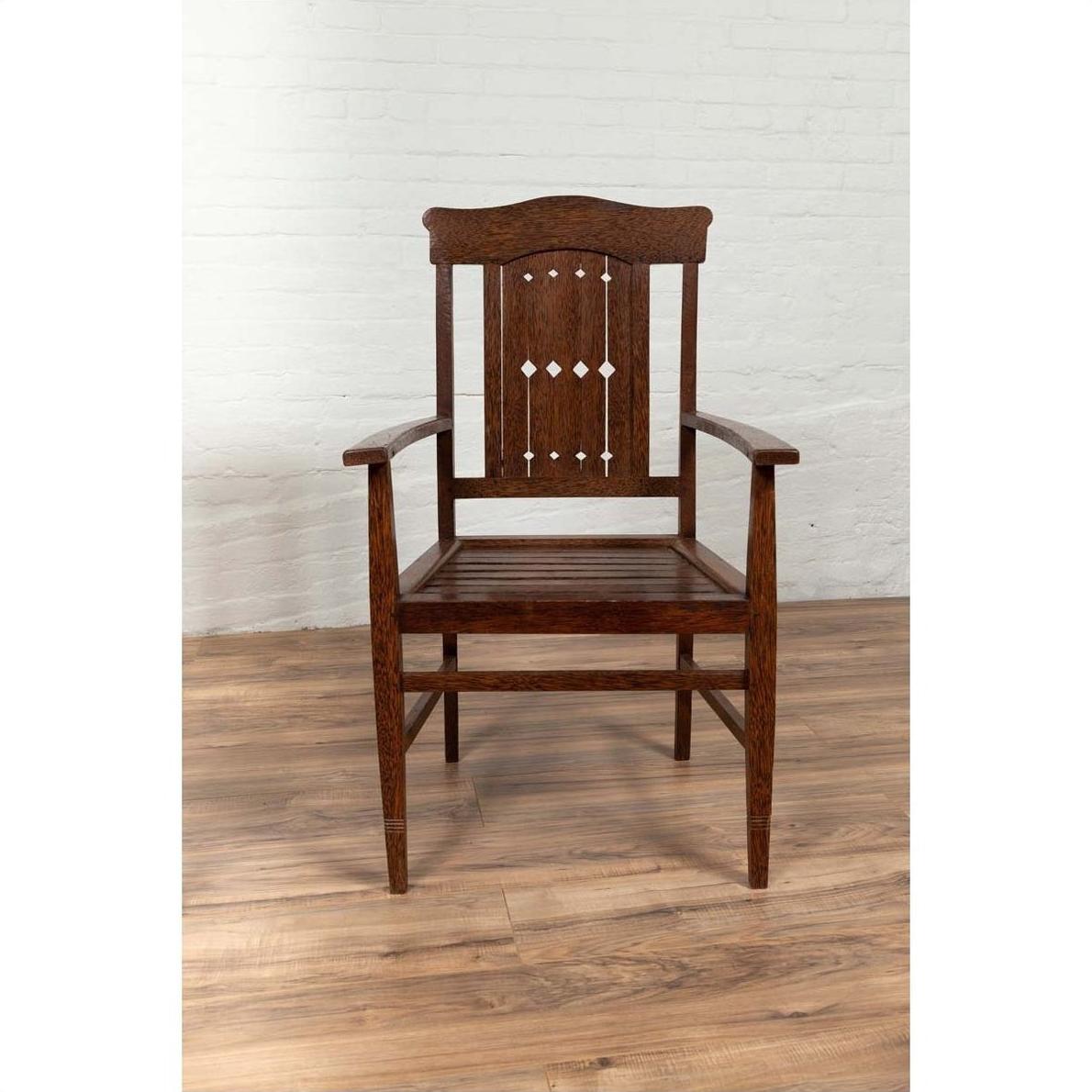 Brown wooden Dutch Colonial armchair with pierced slats, on wood floor next to a white brick wall indoors.