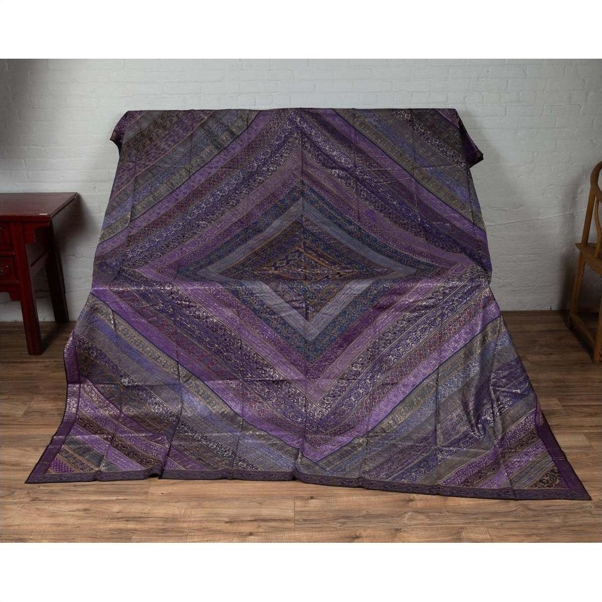 Purple silk fabric with silver and gold accents displayed on a surface next to a wooden floor and white brick wall.