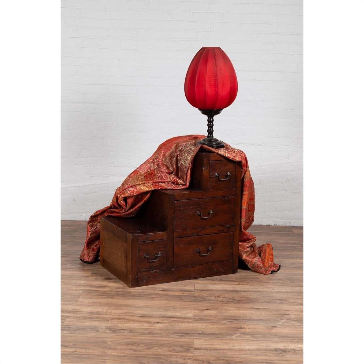 Wooden staircase tansu cabinet with four drawers, red lamp, and patterned cloth on wooden floor indoors.