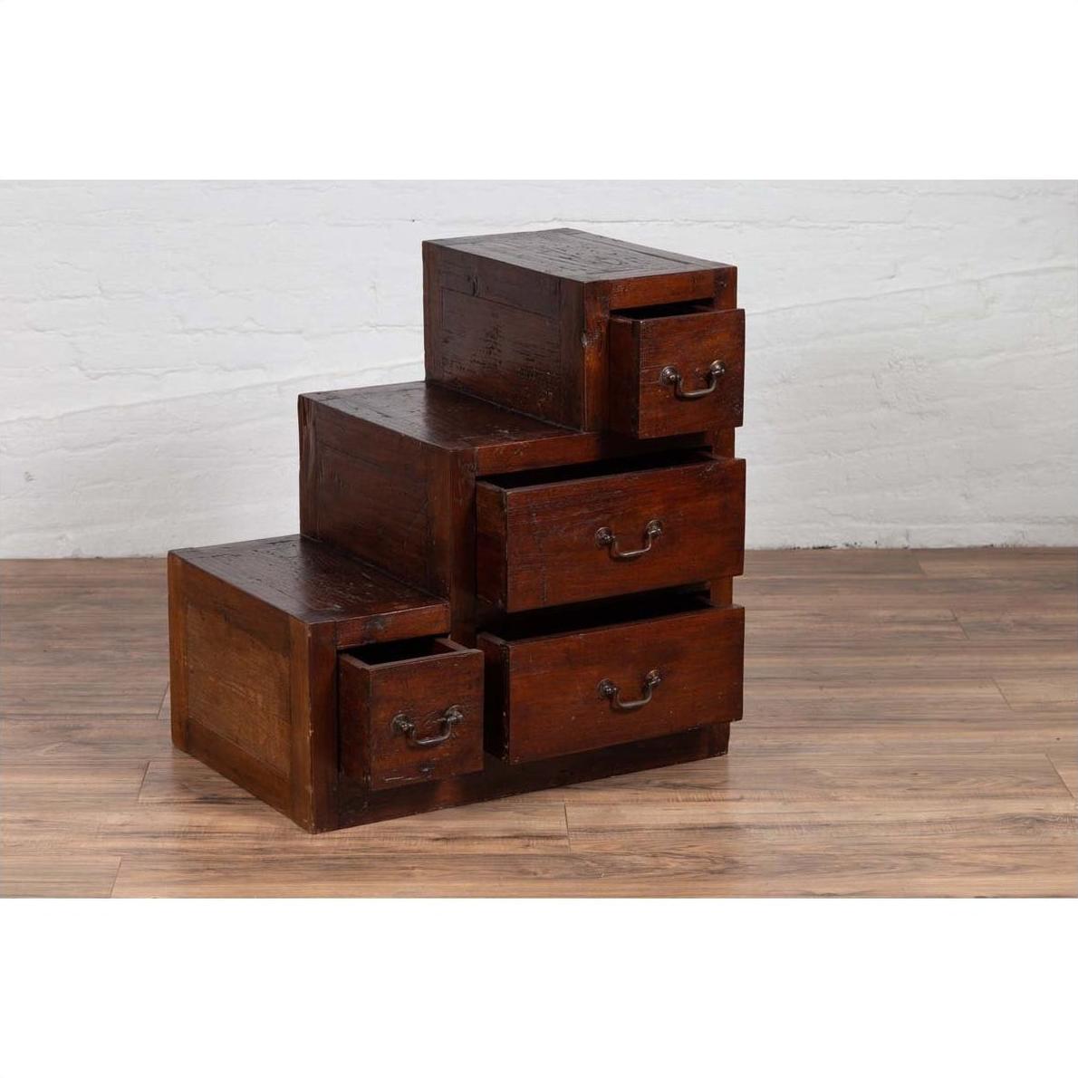 Brown wood Javanese staircase tansu cabinet with four drawers on wooden floor against a white brick wall indoors.