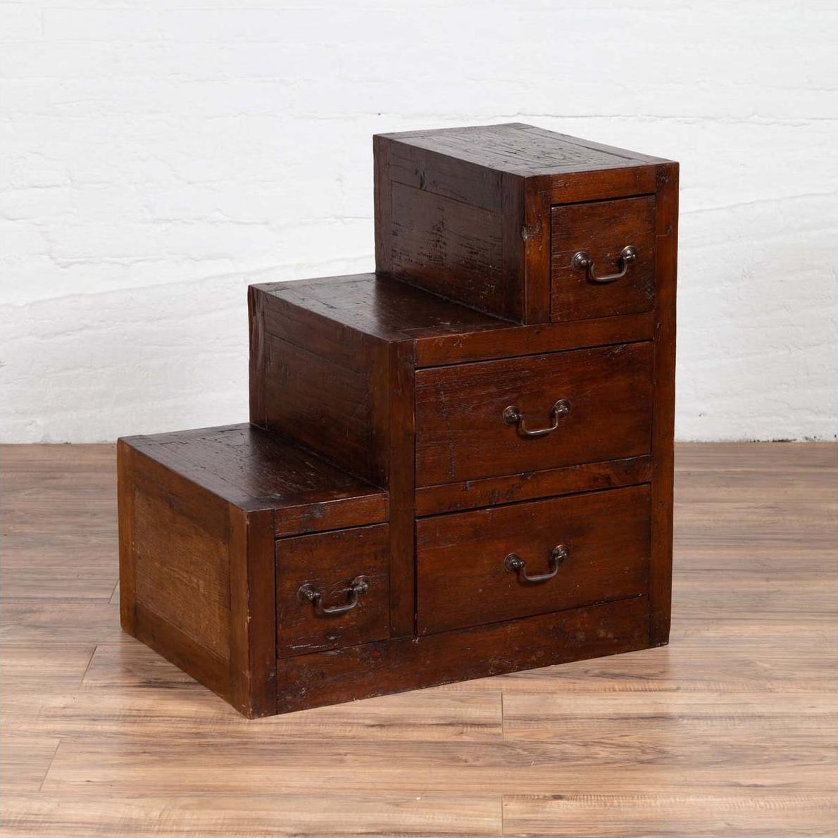 Brown wood staircase tansu cabinet with four drawers, positioned indoors on wooden floor near white brick wall.