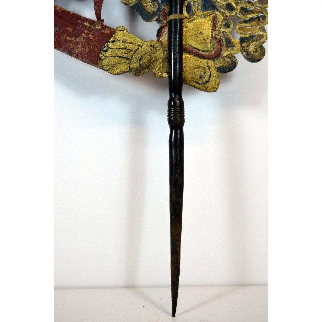 Close-up of tan cowhide Wayang Kulit Balinese shadow puppet held by a gloved hand with a pointed wooden staff.