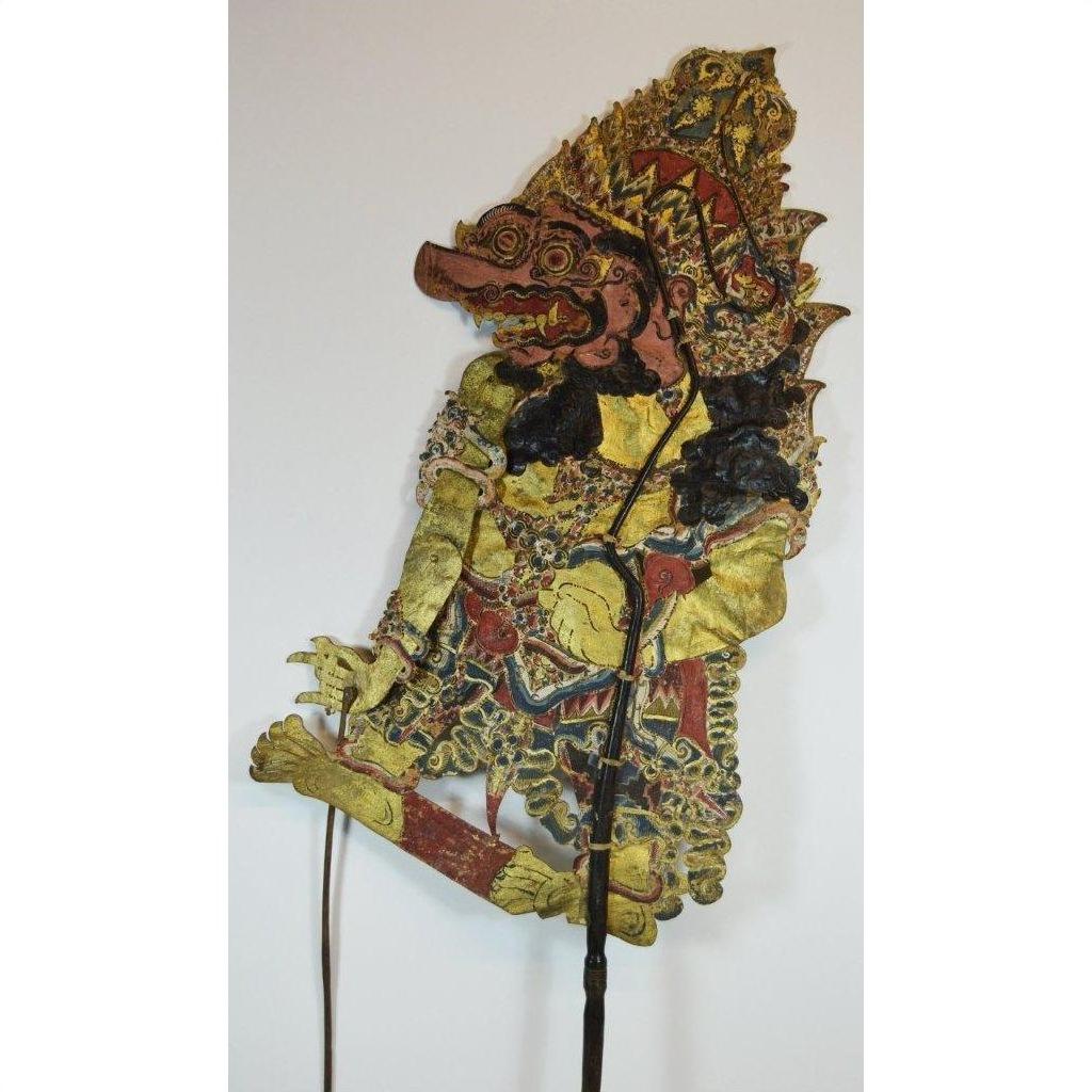 Brown cowhide Wayang Kulit Balinese shadow puppet with ornate costume, large headdress, bold facial features, plain backdrop.
