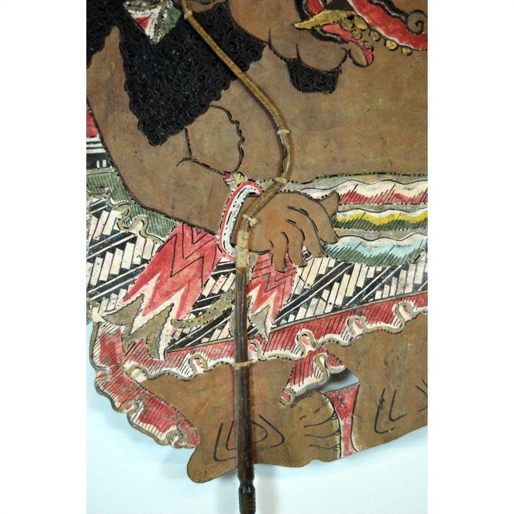 Close-up of painted Balinese cowhide Wayang Kulit shadow puppet with intricate patterns, hand holding wooden stick.