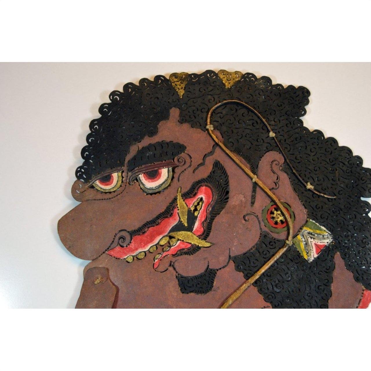 Brown cowhide shadow puppet with curly hair, large red eyes, and ornate earrings, Balinese Wayang Kulit style.