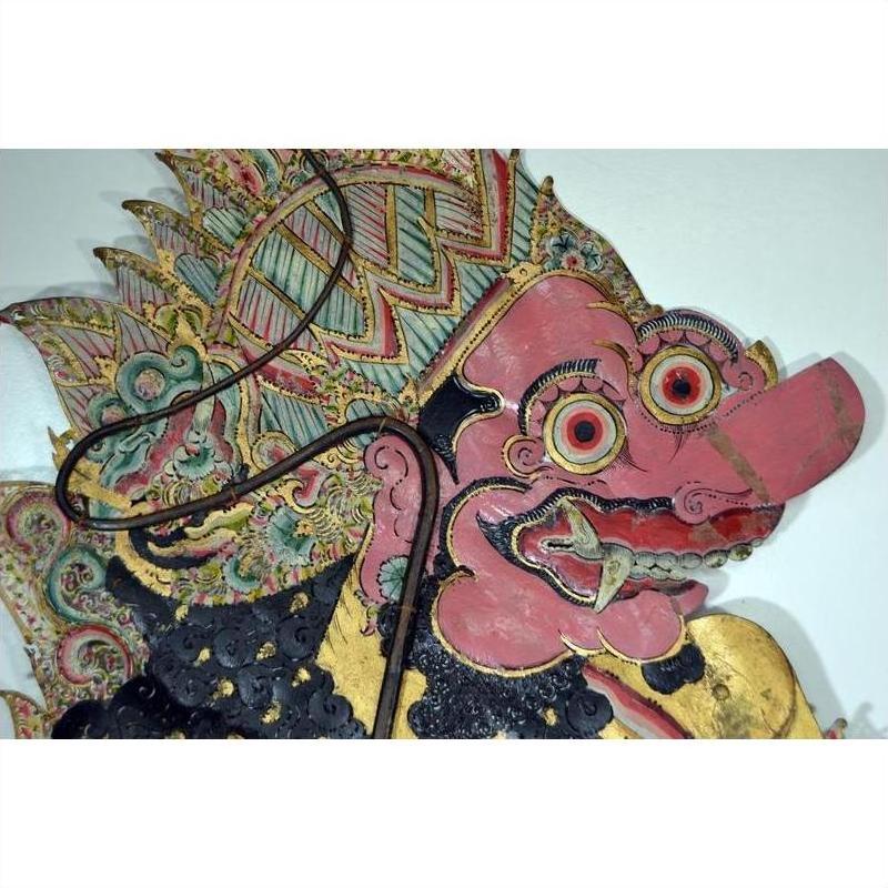 Close-up of a pink-faced Wayang Kulit shadow puppet made from Balinese cowhide, with large eyes and ornate headdress.